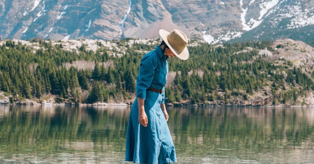 A woman in a blue dress and hat walking on clear rocky shoreline with a mountain backdrop.